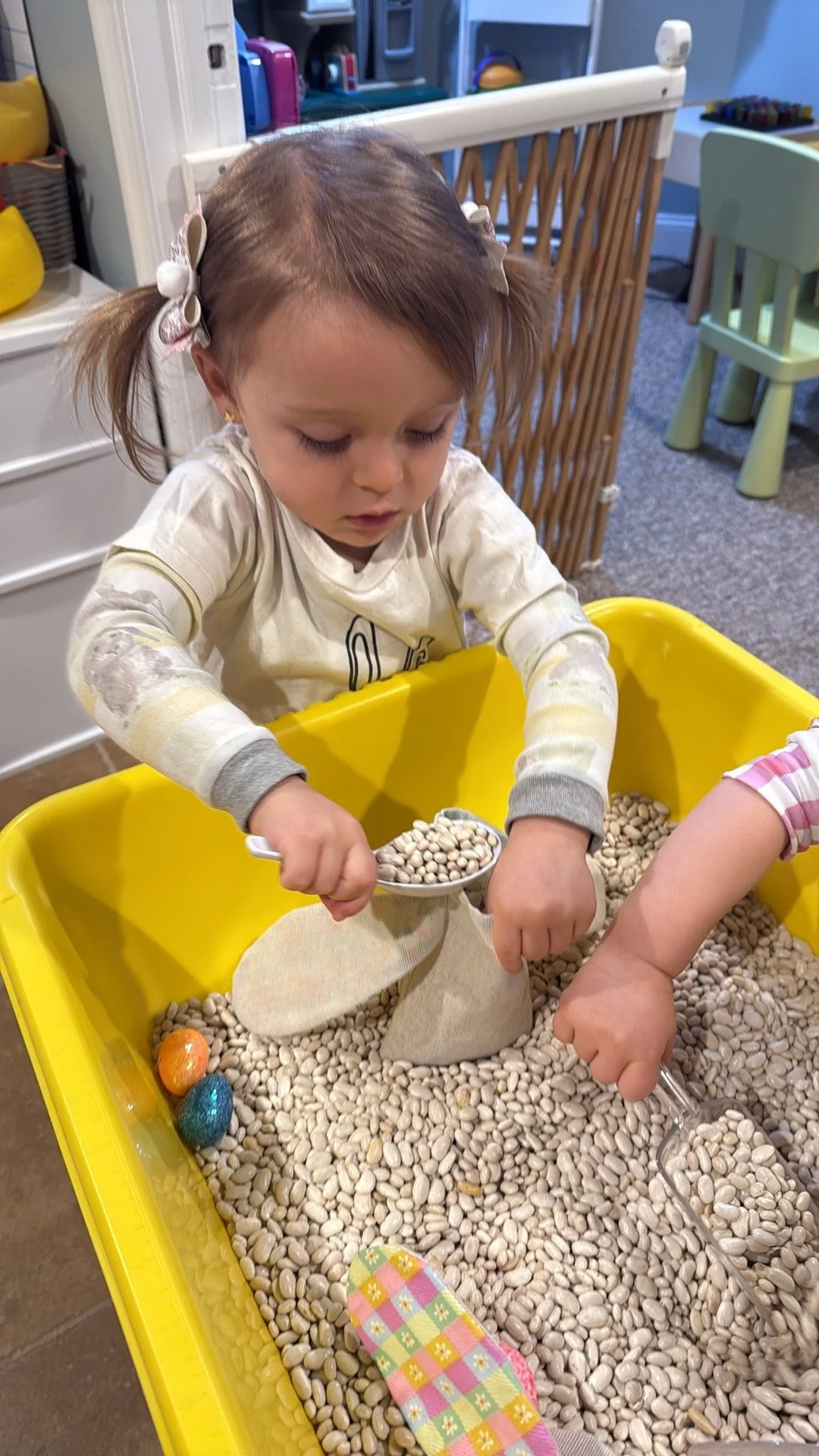 Young girl playing in a sensory bin filled with white beans, scooping beans with a metal scooper, with Easter eggs visible in the bin.