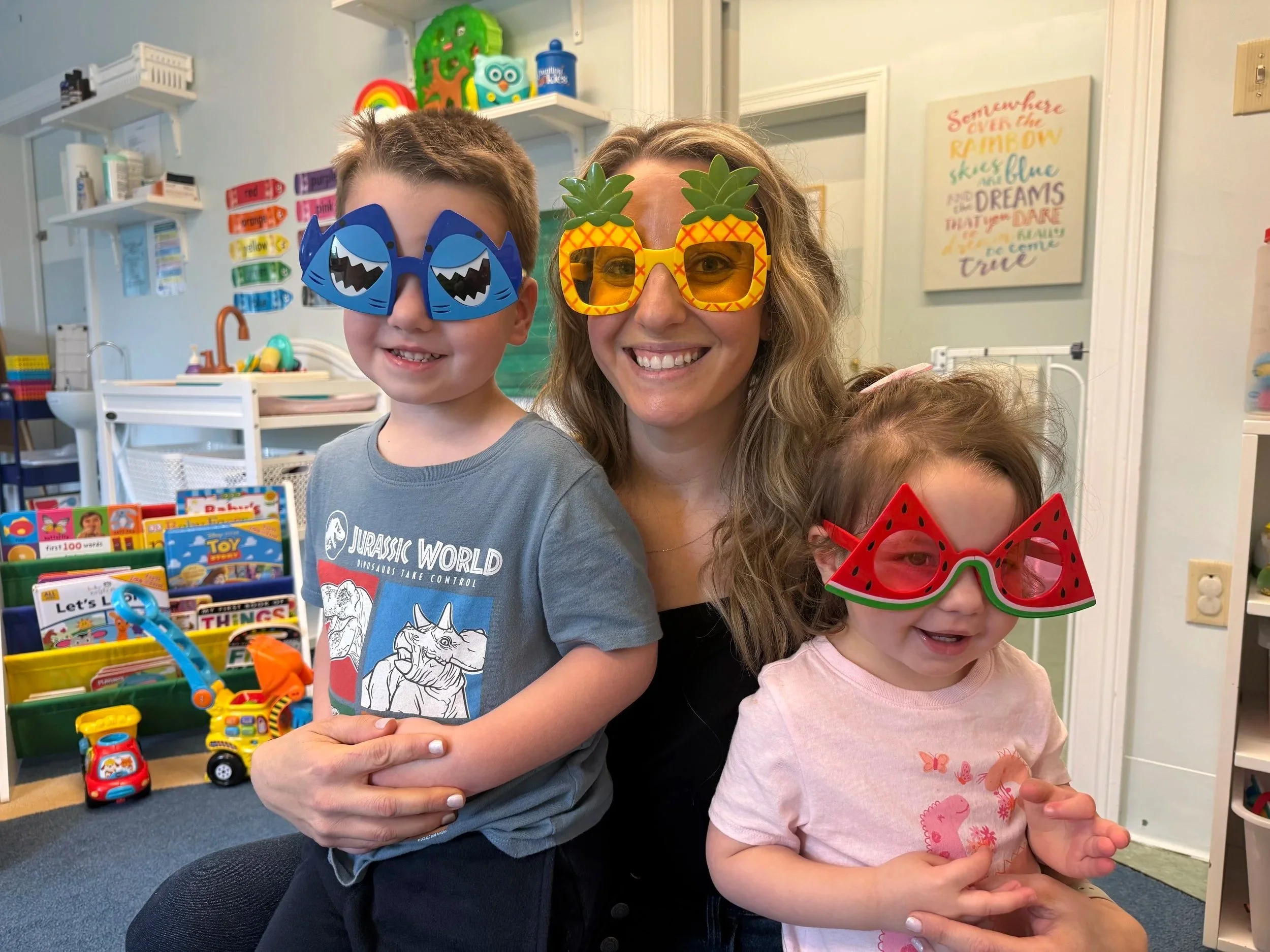 A woman and two children wearing colorful novelty glasses, smiling at the camera in a play or classroom setting with toys and educational materials in the background.