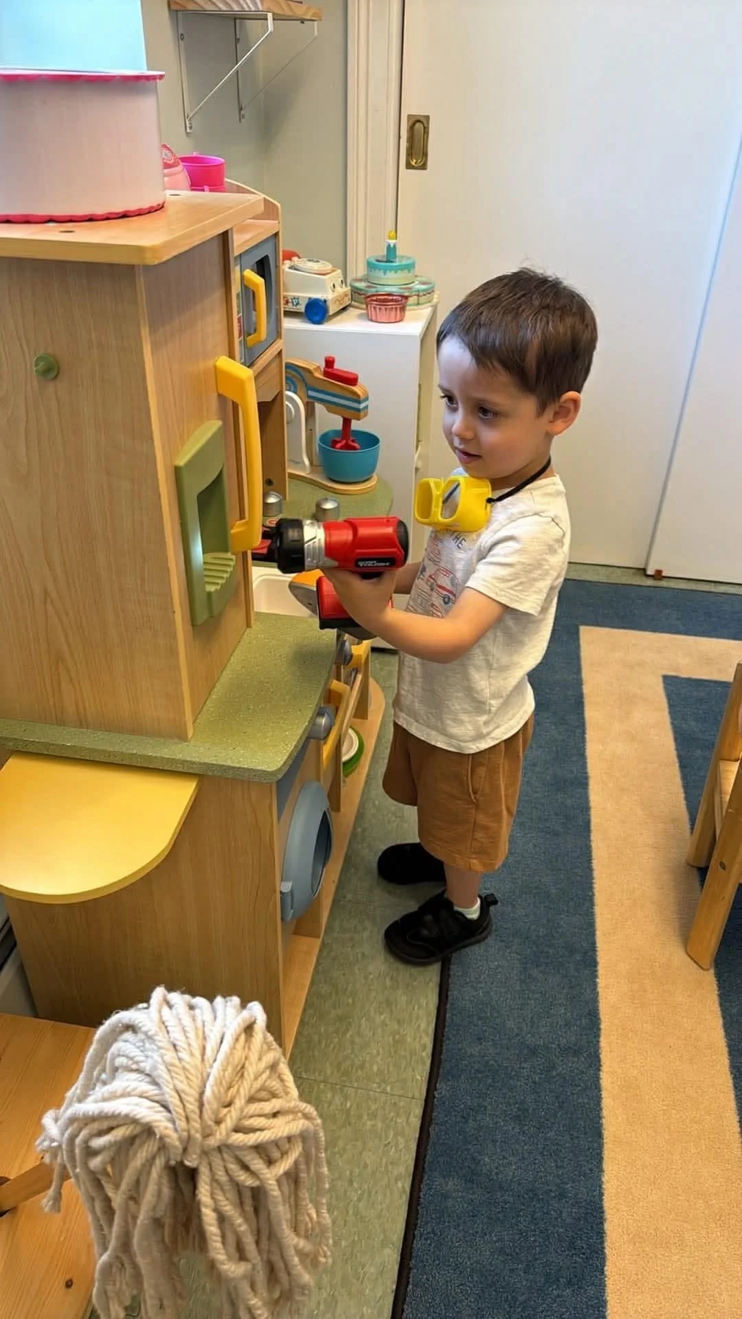 A young boy wearing a white T-shirt, brown shorts, and black shoes is playing with a red toy cordless drill in a play kitchen area. The toy is held to a wooden cupboard with colorful handles. There is a ball of thick beige yarn hanging from a wooden 
