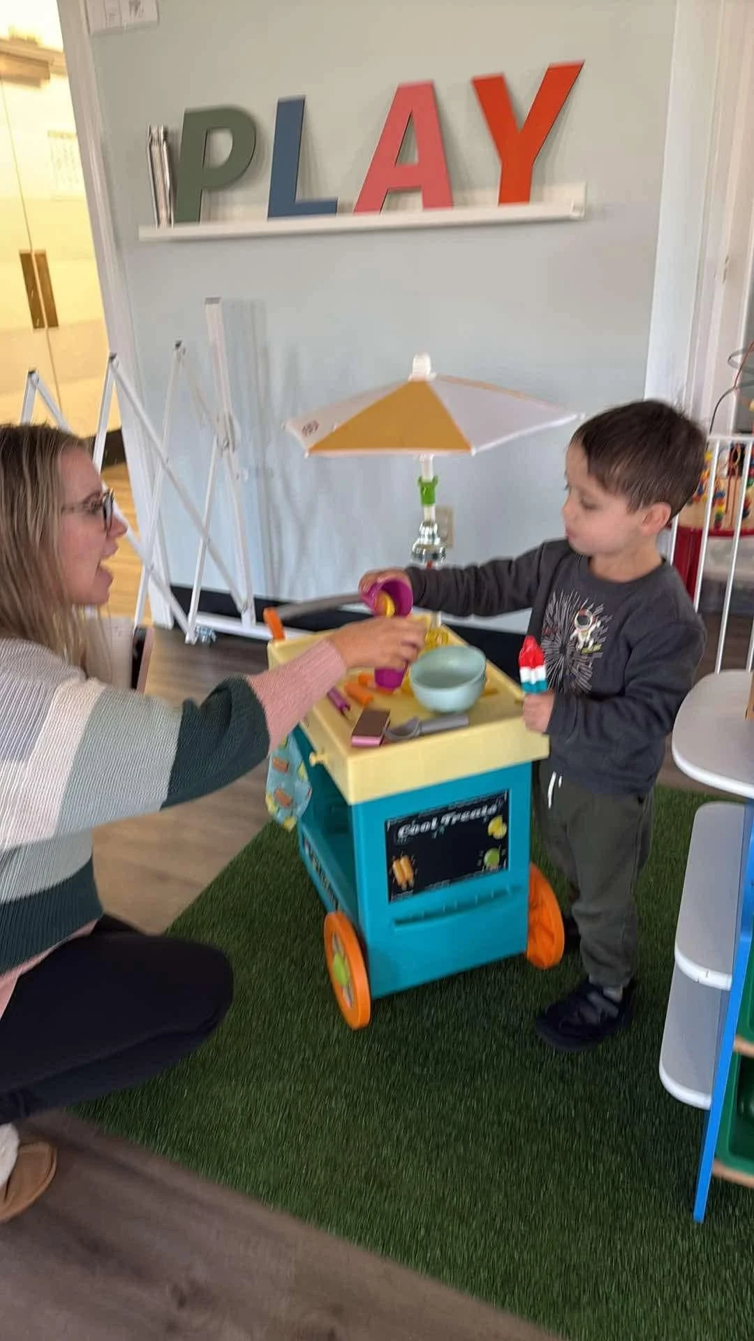 Child playing with a toy food cart while a woman interacts with him, in a room with a sign that says 'PLAY' on the wall.
