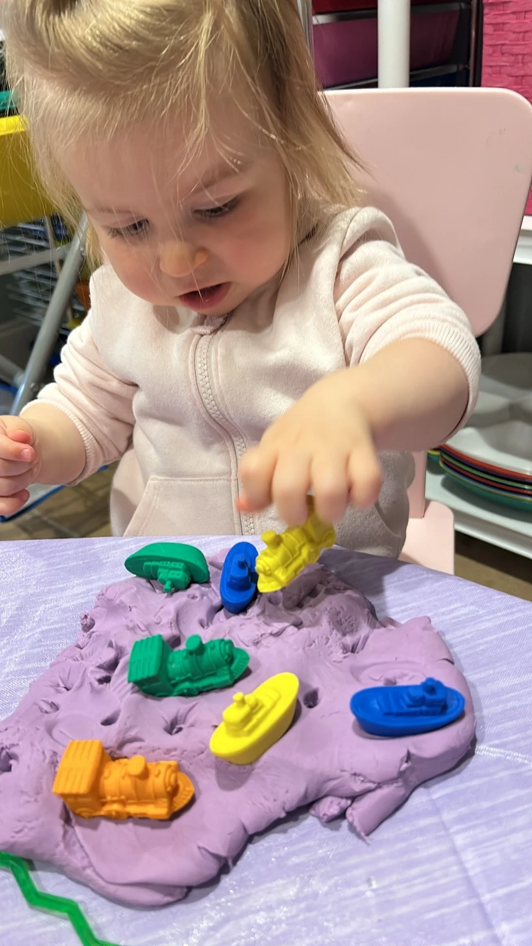 A young girl playing with colorful toy boats on a purple mound of slime at a table.