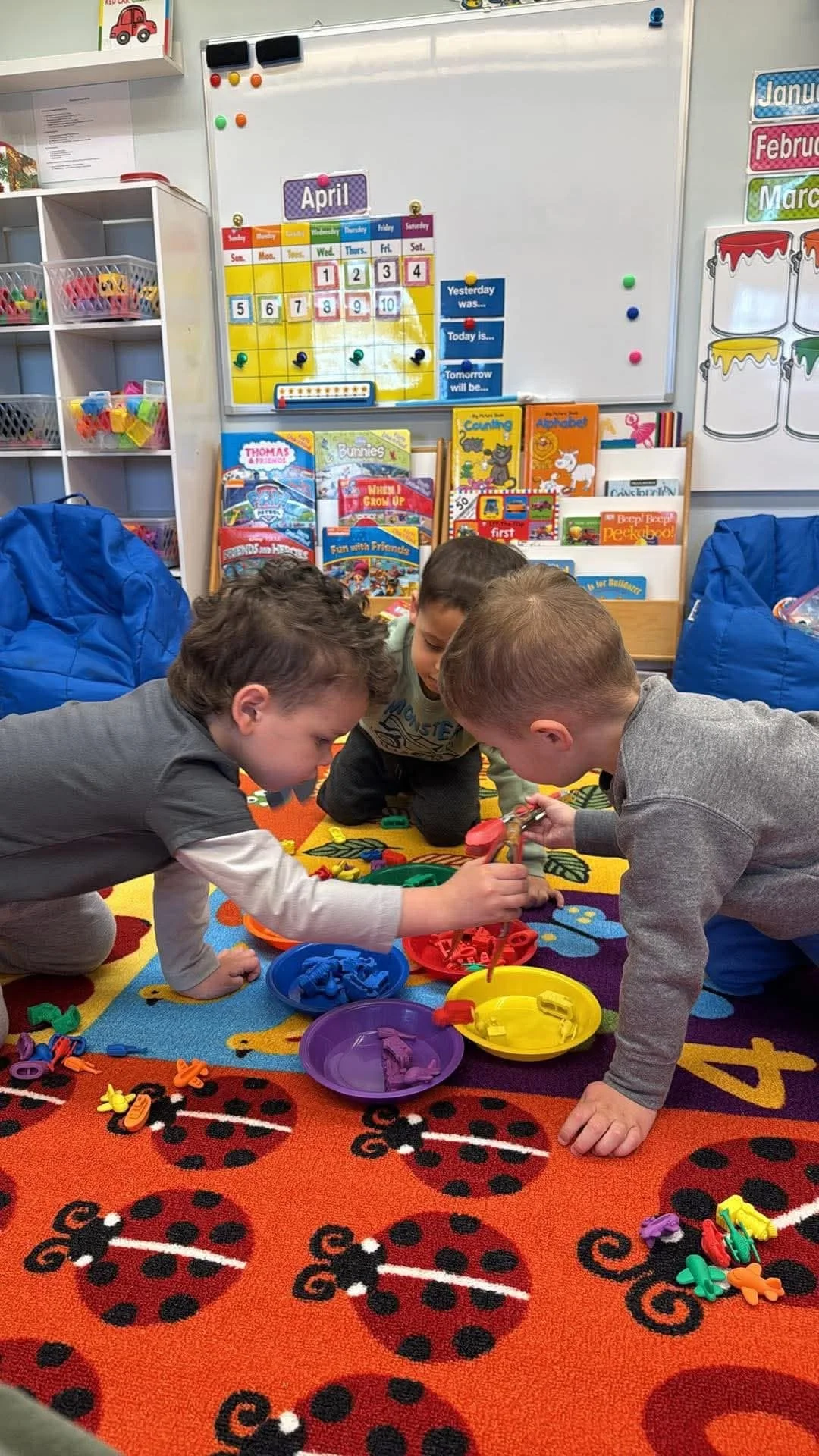 Three young boys are playing with colorful rubber bands and clips on a playful ladybug-patterned carpet in a classroom. Behind them, there are bookshelves filled with children's books, a whiteboard, and a display of months and days.