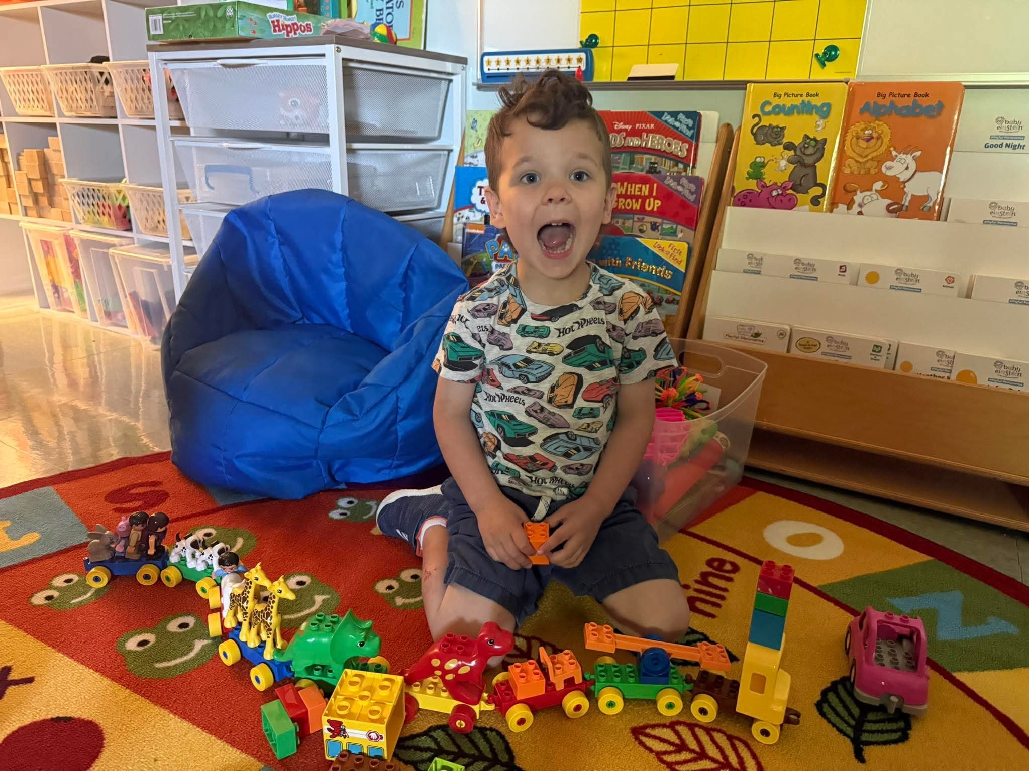 A young boy sitting on a colorful animal-themed rug in a classroom play area, surrounded by various toy animals, blocks, and building toys, with educational books and storage shelves in the background.