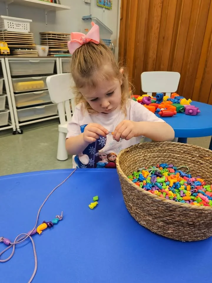 A young girl with a pink bow in her hair is sitting at a blue table, stringing beads to create a necklace or bracelet, with a large basket of colorful plastic beads and some completed jewelry on the table.