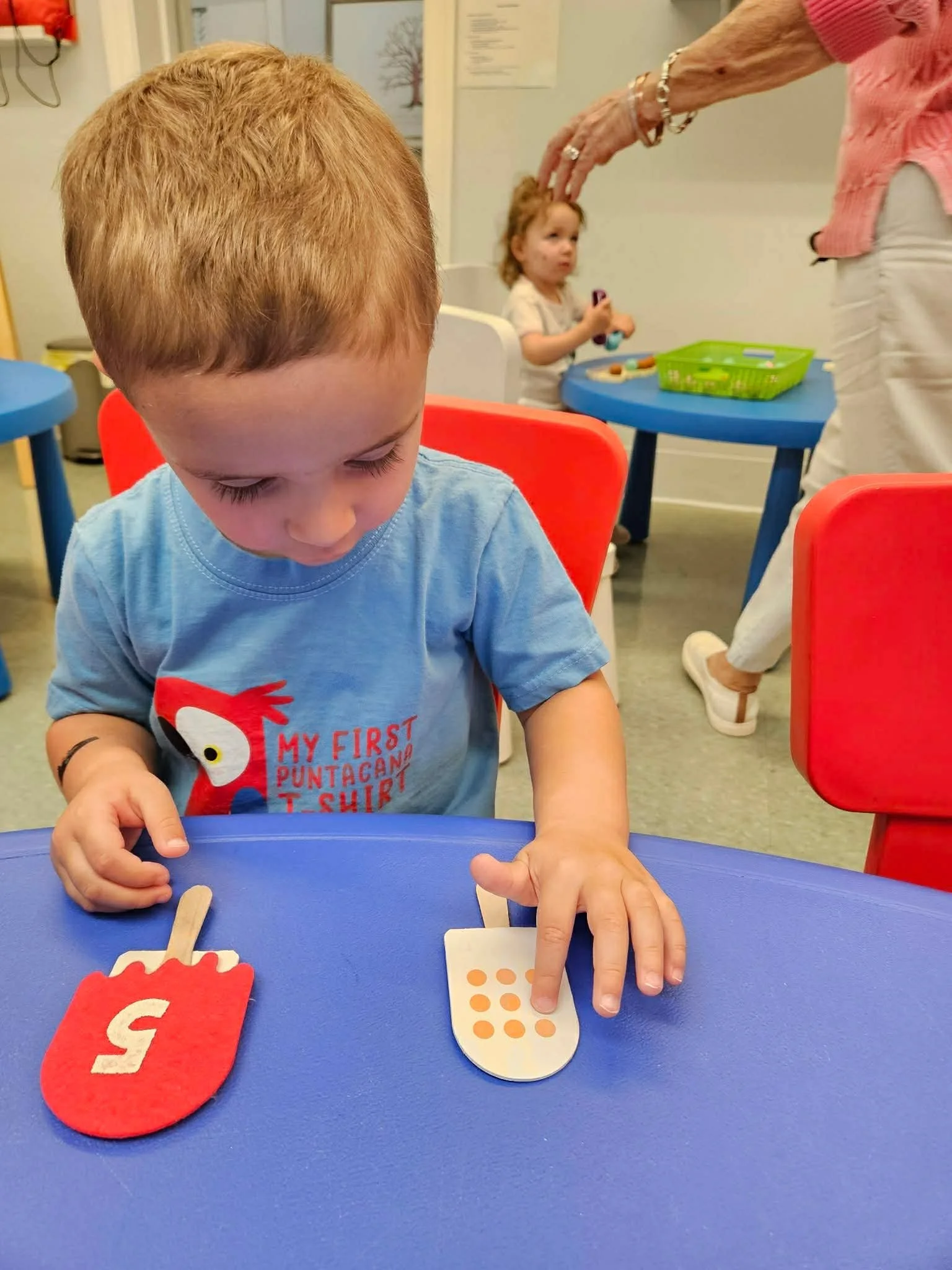 Child with red hair playing with educational toys at a blue table, a woman reaching in the background, and another child sitting at a table