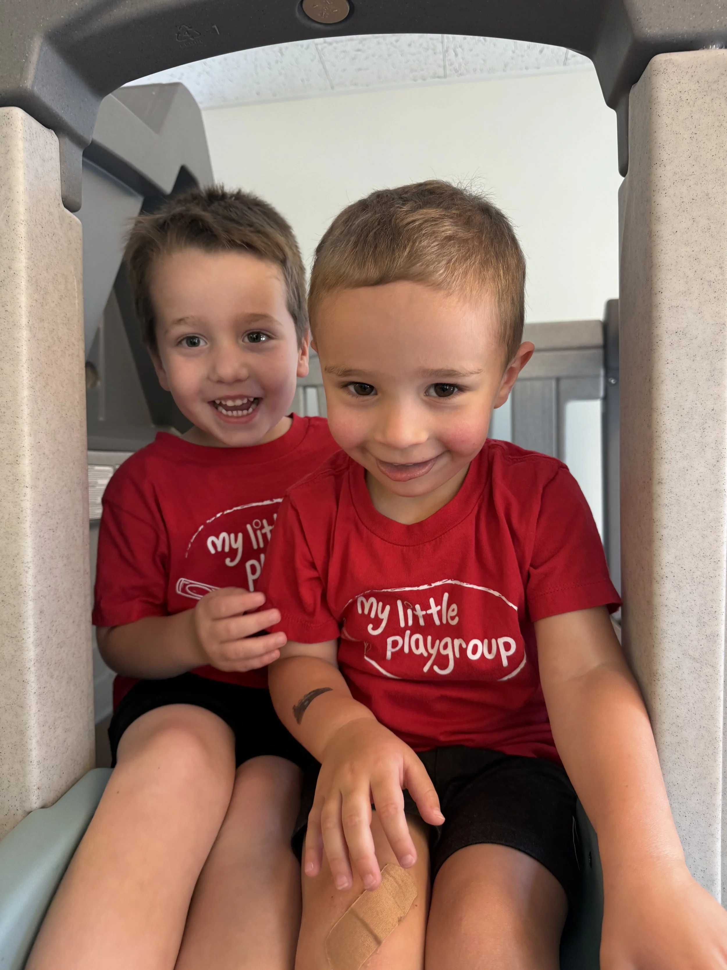 Two young boys with short brown hair sitting under a table, both wearing red shirts with white text that reads 'my little playgroup.' One boy is smiling with mouth open, the other is looking down with a slight smile. 