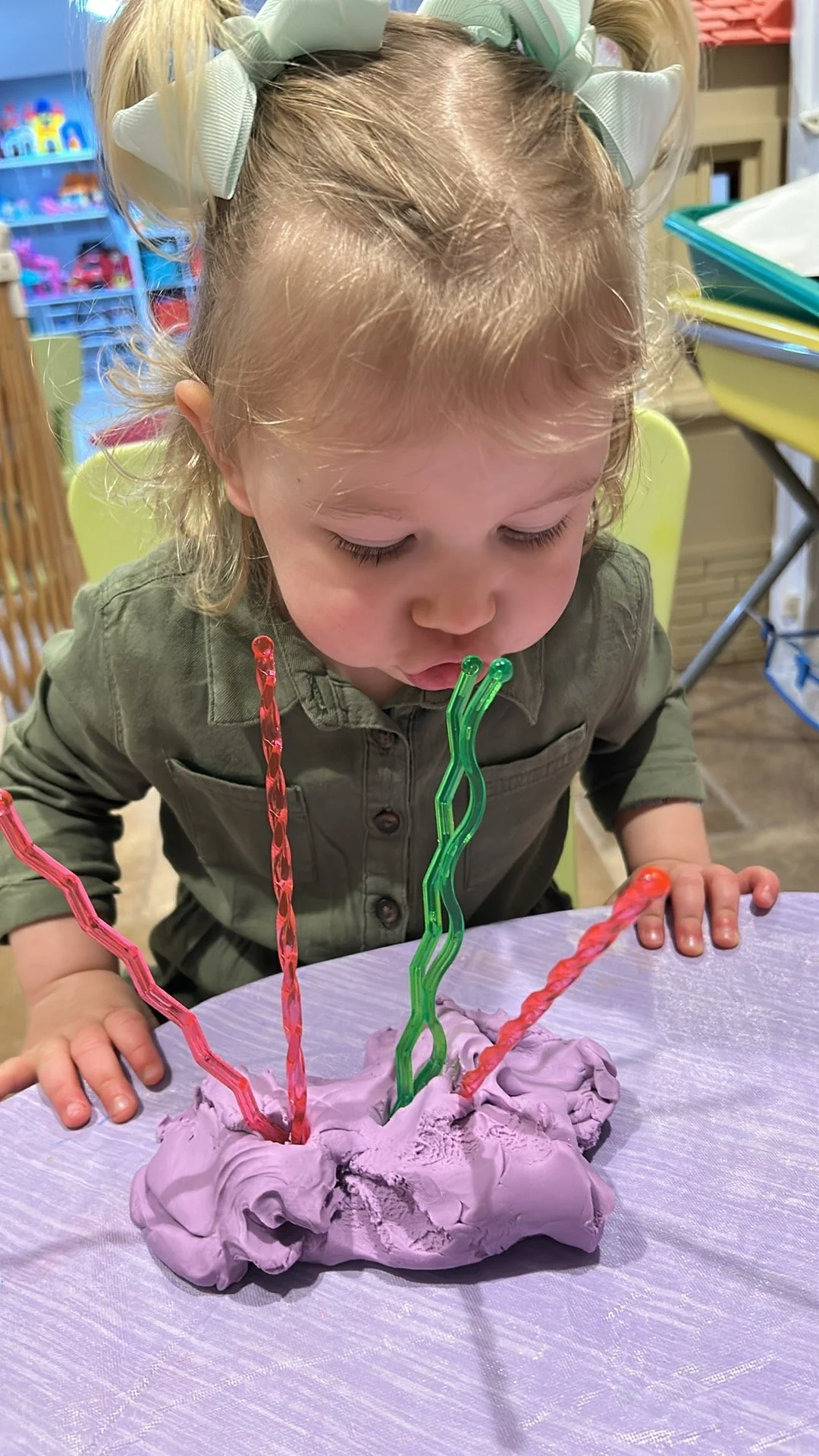 A young girl with blonde hair tied with large light green bows, blowing out candles on a purple birthday cake with colorful candles, in a decorated indoor setting.