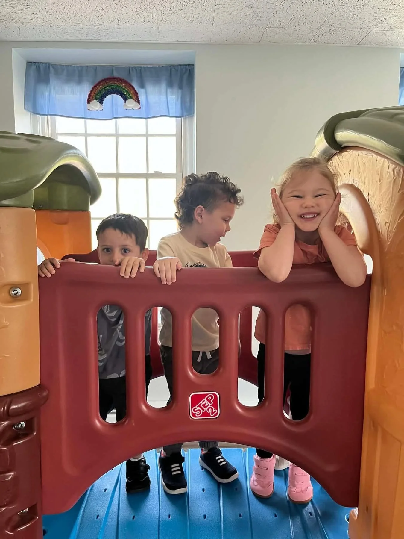 Three children standing inside a colorful play structure with a red barrier and a rainbow decoration on a window behind them, in a room with white walls and blue flooring.