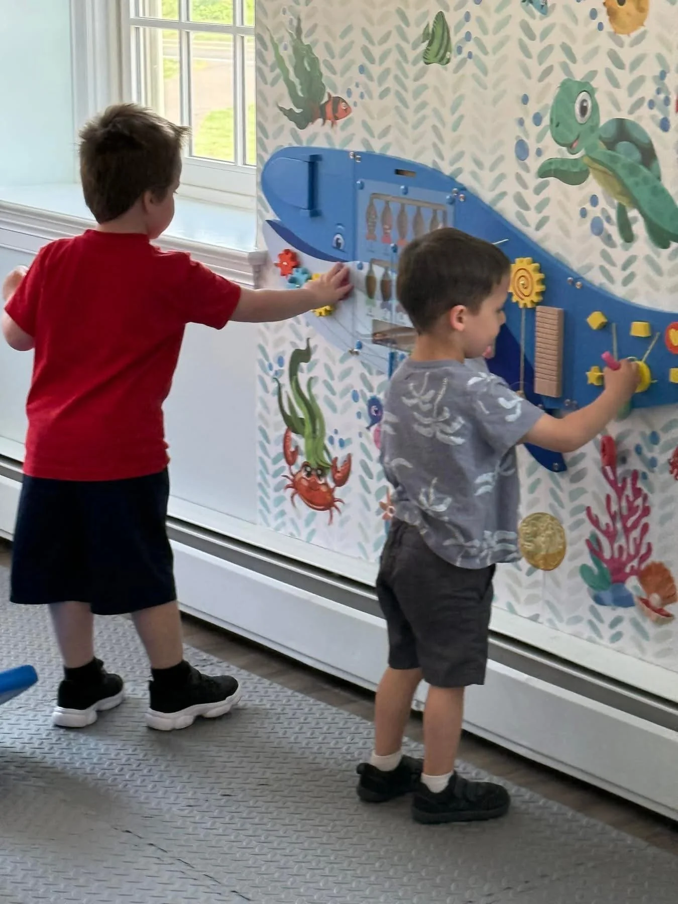 Two young boys playing with an interactive wall display featuring nautical and ocean-themed artwork of fish, crabs, and an underwater scene.
