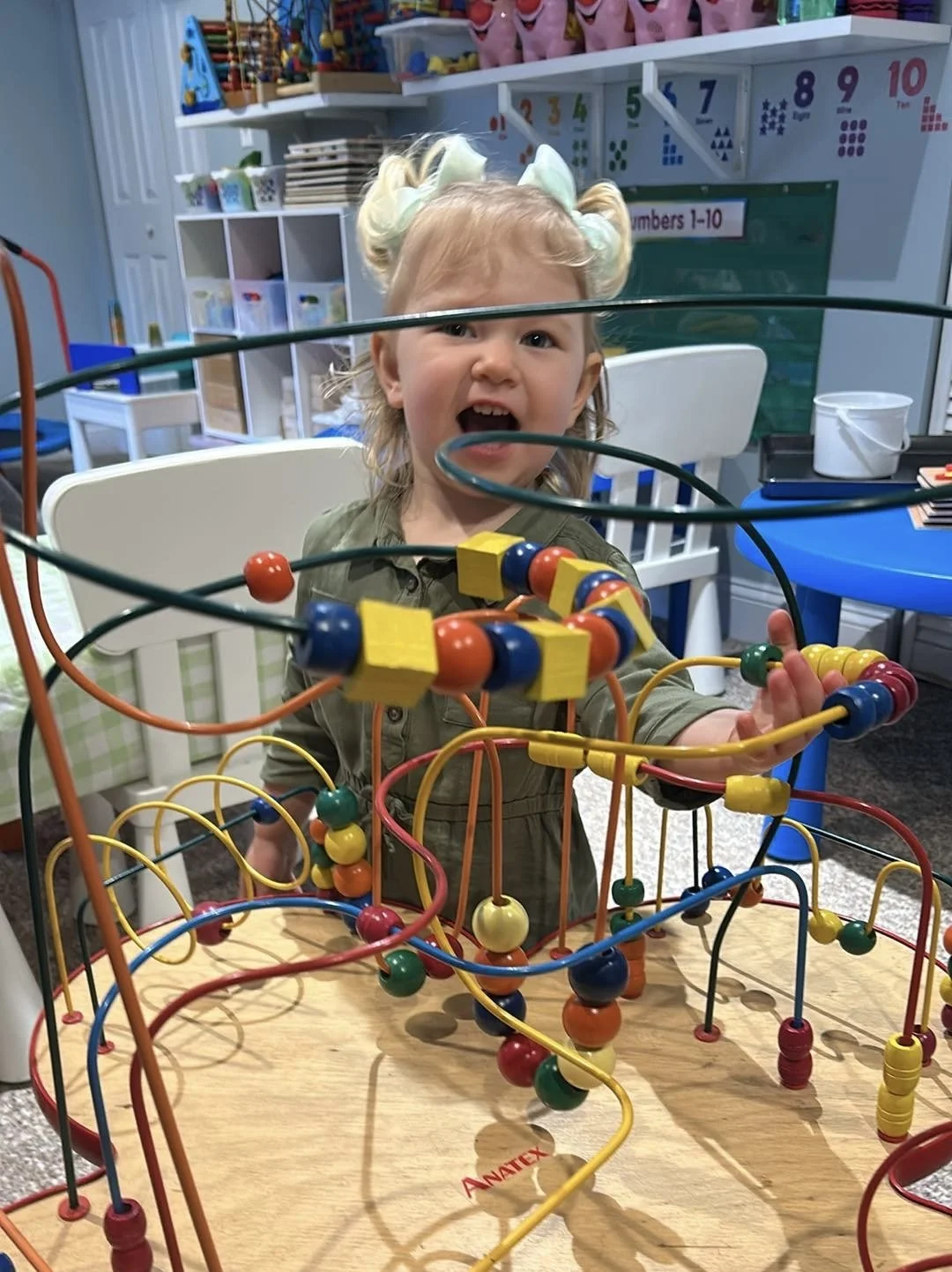 A young girl with blonde hair in pigtails and a green outfit, smiling and playing with a colorful bead maze toy inside a classroom or playroom.