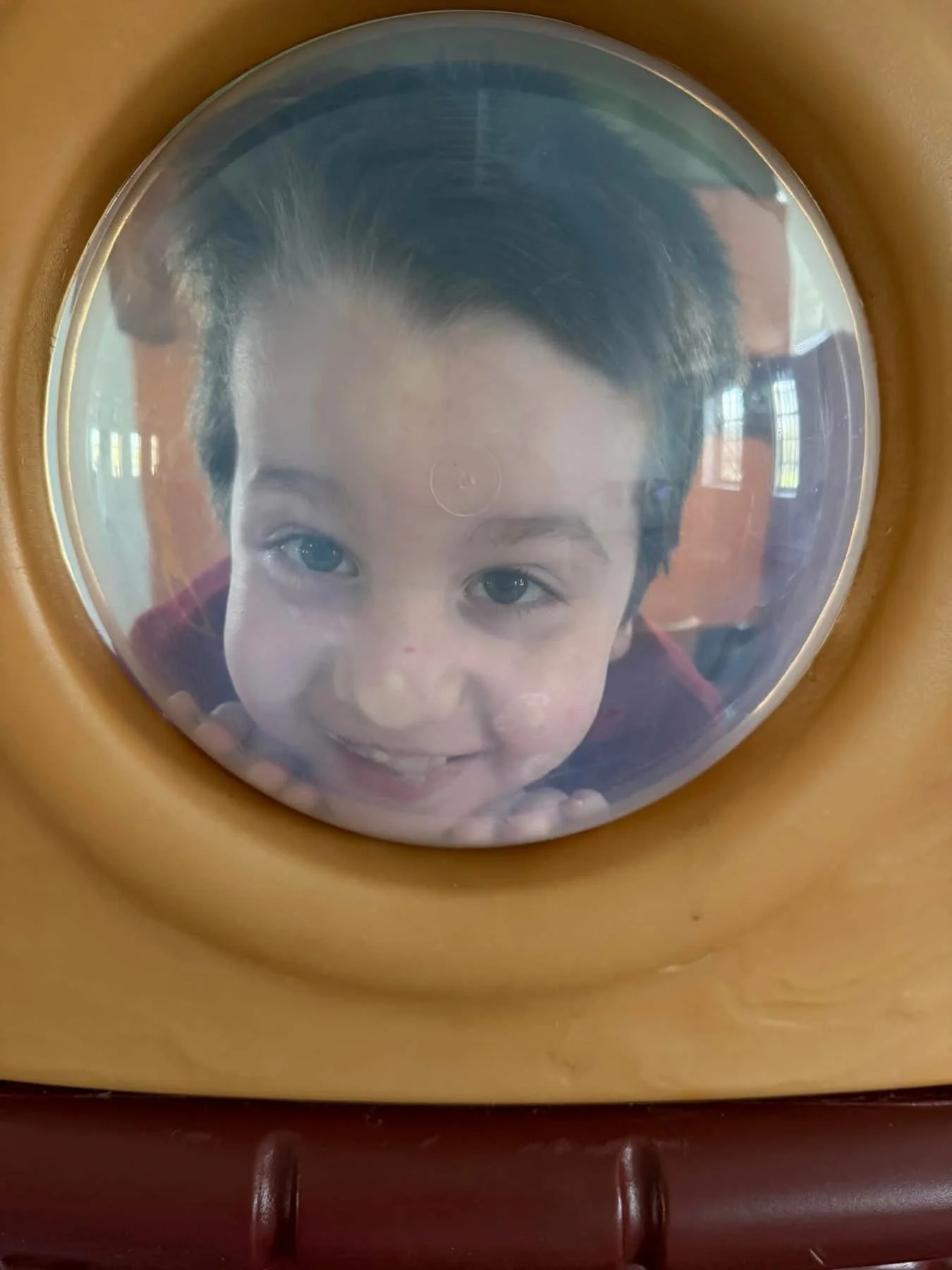 Child looking through a circular window in a playground structure with a happy expression.