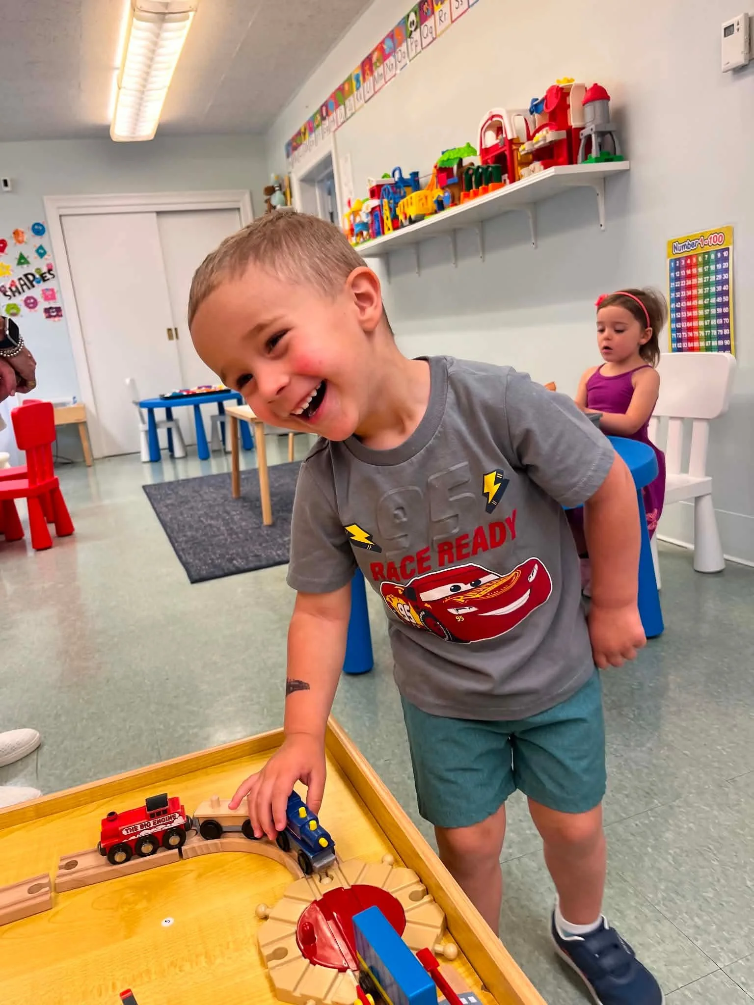 A young boy in a gray t-shirt and shorts playing with toy train set in a classroom with colorful decorations, including a girl sitting in the background, a wall with alphabet and number charts, and shelves with toys.