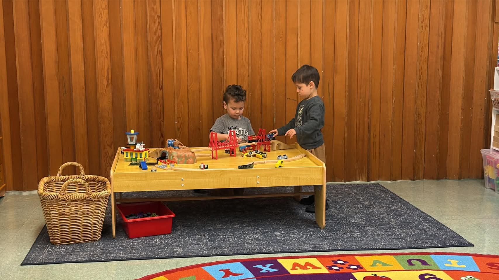 Two young boys playing with toy cars and a train set at a wooden table in a playroom. Wooden wall background, a basket on the left, and a colorful alphabet rug on the floor.