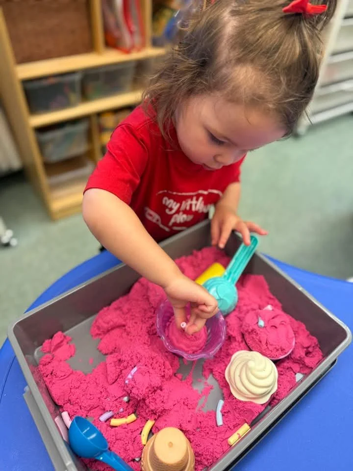 A young girl playing with pink kinetic sand in a plastic tray, using various colorful tools and molds, in a classroom setting.