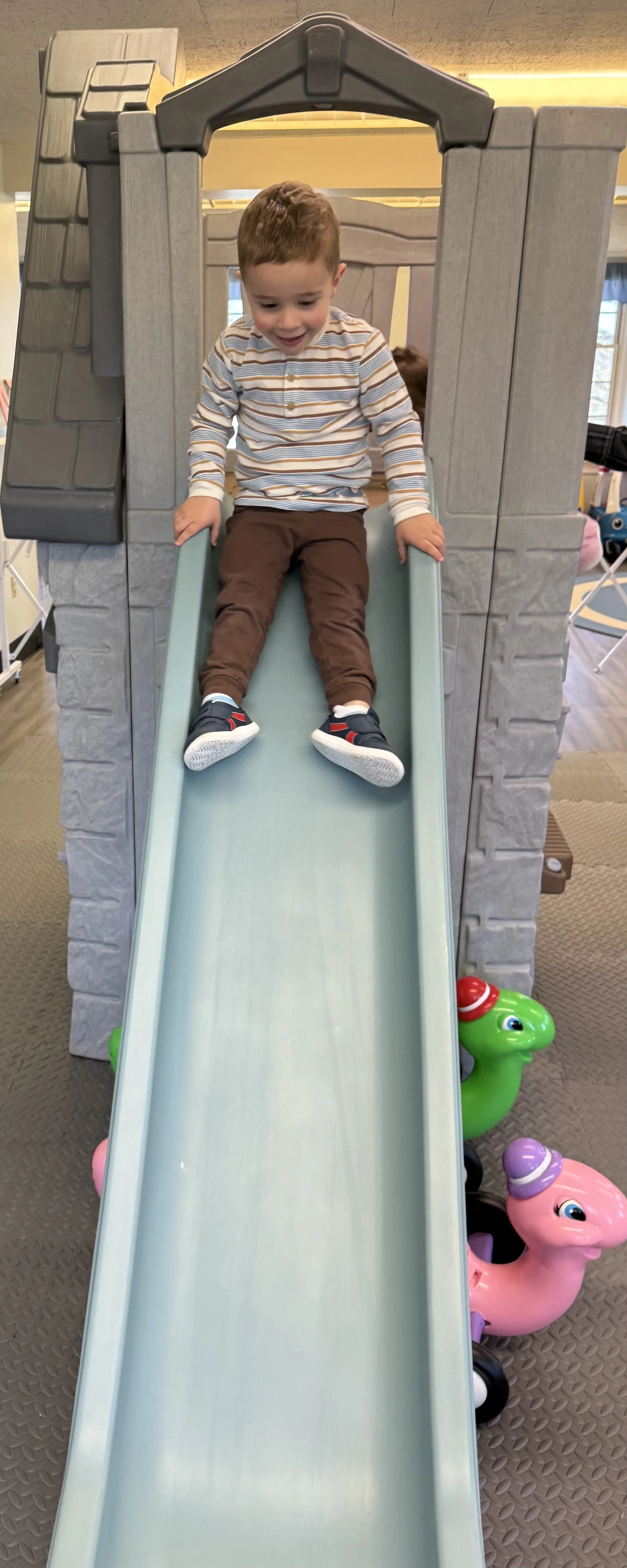 A young boy with short brown hair is sitting at the top of a small plastic slide, smiling. The slide is part of a playhouse structure with a faux stone facade. There are two colorful ride-on toys shaped like animals at the base of the slide, one gree