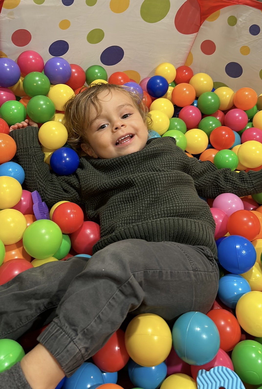A young boy is lying on his back in a colorful ball pit filled with plastic balls of various colors including yellow, red, green, blue, pink, and orange. He is smiling and looks happy.