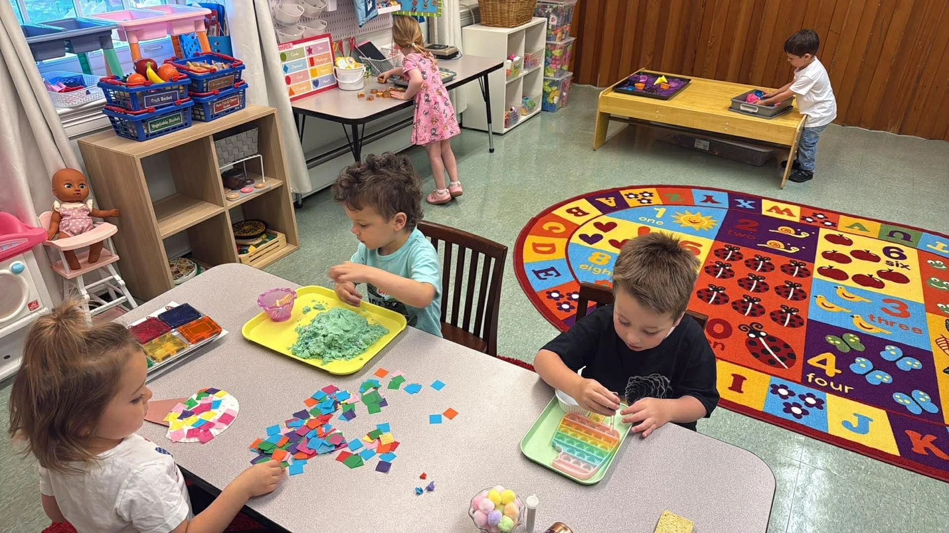 Children engaged in arts and crafts at a classroom table decorated with colorful confetti and craft supplies. In the background, a play area with a brightly colored rug featuring numbers and animals, and shelves with toys and educational materials.