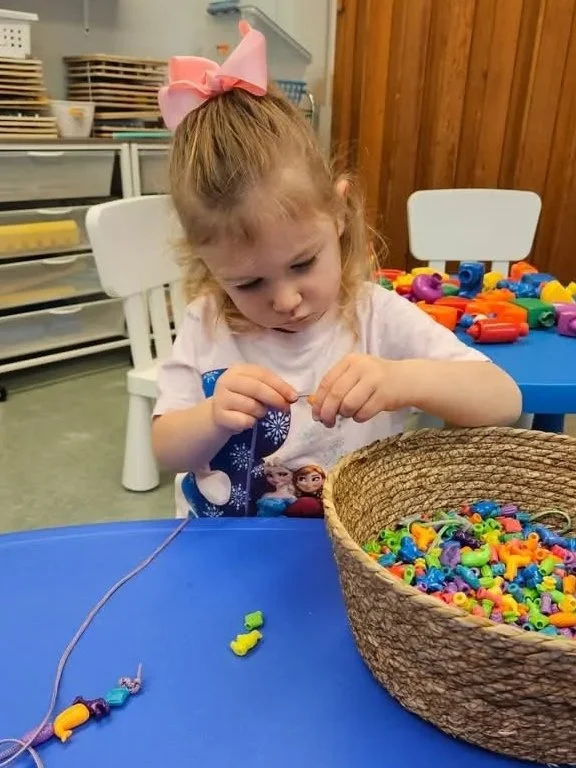 Young girl with a pink bow in her hair, wearing a Disney Frozen shirt, sitting at a blue table, stringing colorful beads from a large woven basket. Rearranged beads and toys are visible on her table and in the background.