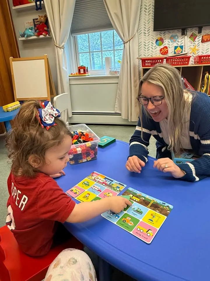 A young girl with a bow in her hair is playing a matching game with a woman at a blue table in a classroom. The girl is pointing at a card while the woman is smiling and engaging with her. The background shows a window with curtains, colorful artwork