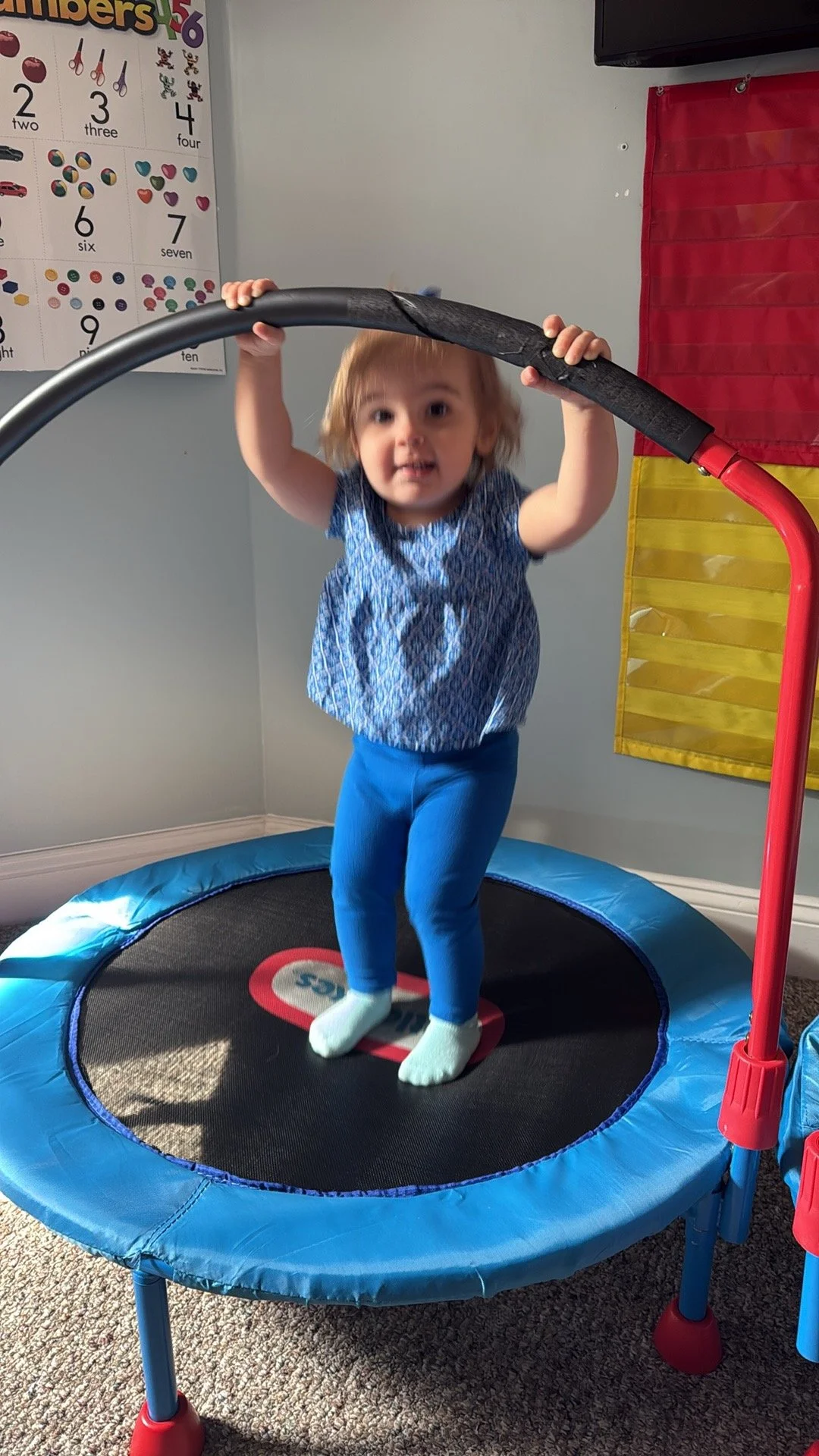 Young girl jumping on a small trampoline indoors, holding onto the safety bar, in a room with educational posters and toys.