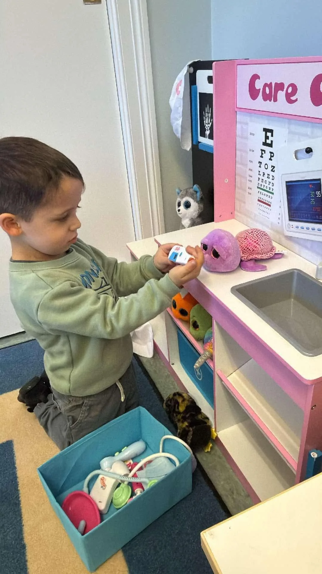A young boy kneeling on the floor next to a pretend medical playset with plush toys, a doll, and toy medical equipment. The playset resembles a doctor's office with a sink, eye chart, and monitor. The boy is holding a toy medical instrument and looki