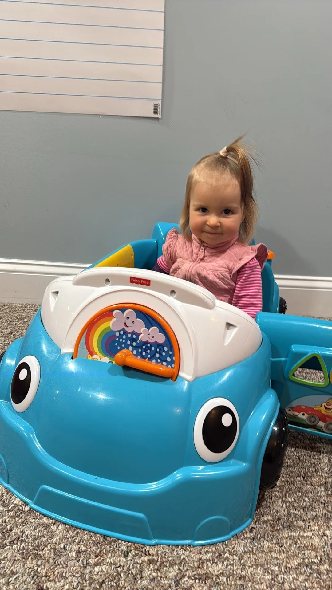 A toddler girl with a ponytail sitting in a blue toy car with a rainbow and cloud design on the front.