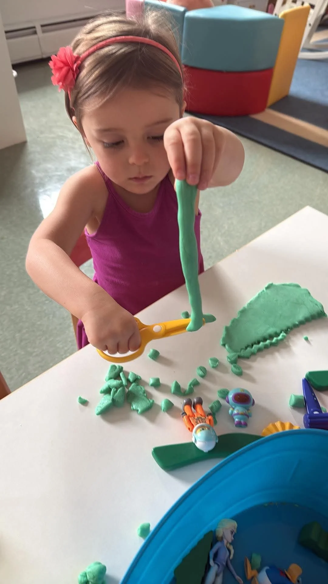 A young girl with a pink headband and purple tank top playing with green slime and small toys at a table.
