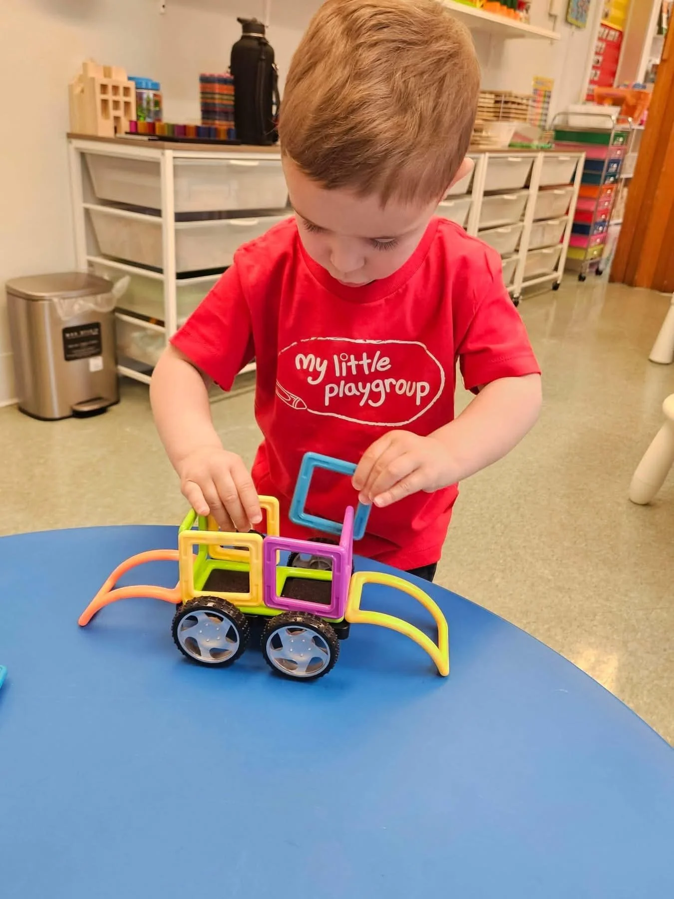 A young boy with light brown hair playing with a colorful magnetic building toy on a blue table in a classroom or play area. He is wearing a red T-shirt that reads "my little playgroup." In the background, there are shelves with toys, books, and a tr