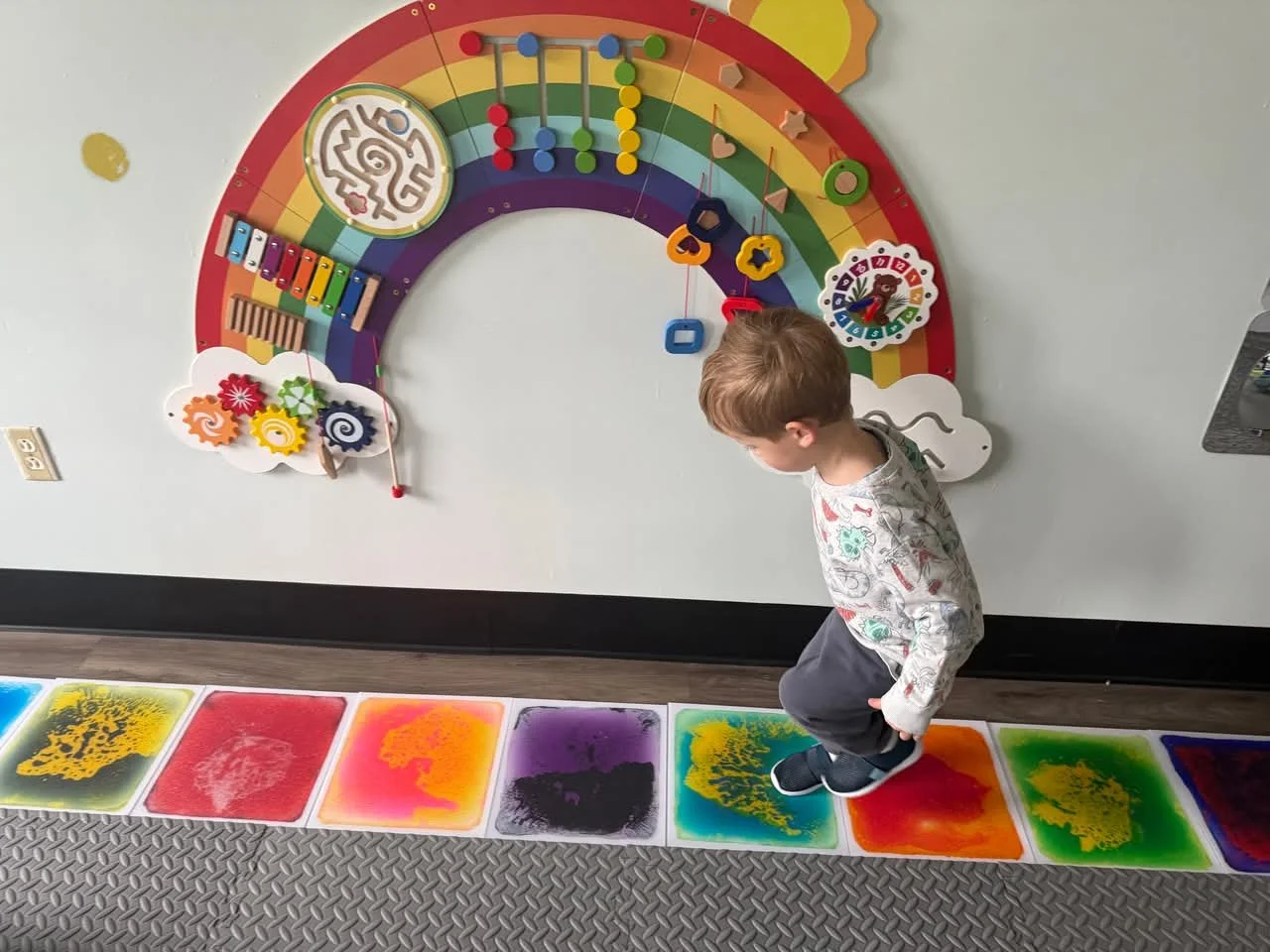 A young boy standing on a colorful splash pad in front of a Wall Wheel toy with various gears, colors, and shapes on a rainbow-themed background.
