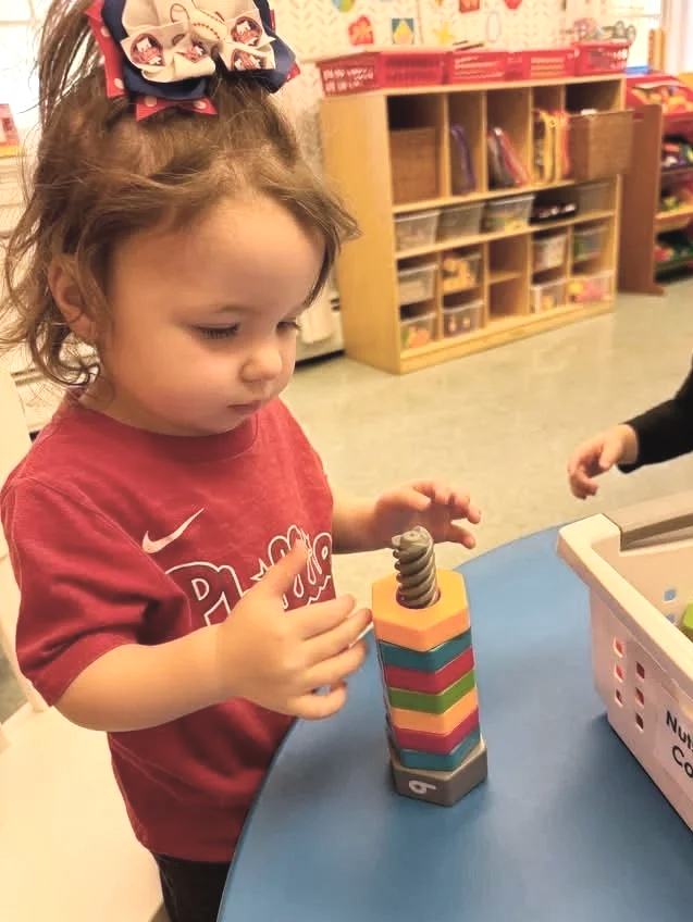 A young girl with curly hair in a ponytail adorned with a bow featuring cartoon faces, playing with a stacking toy in a classroom setting.