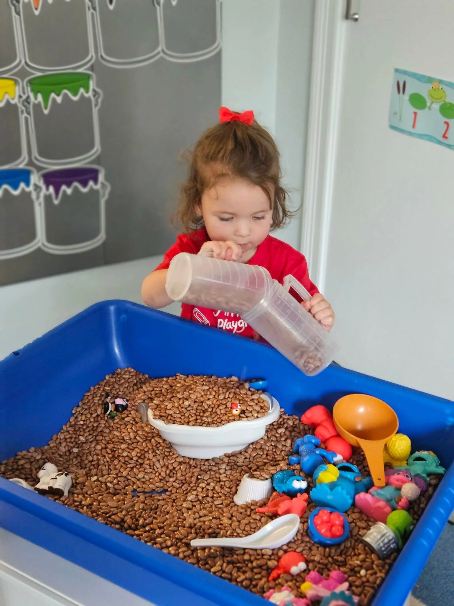 Child with a red bow playing in a sensory bin filled with brown beans and small toys, including animal figures and plastic eggs.