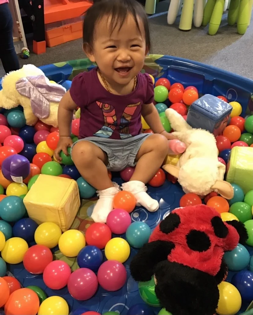 Young child sitting in a ball pit filled with colorful plastic balls and stuffed animals, smiling and enjoying playtime.