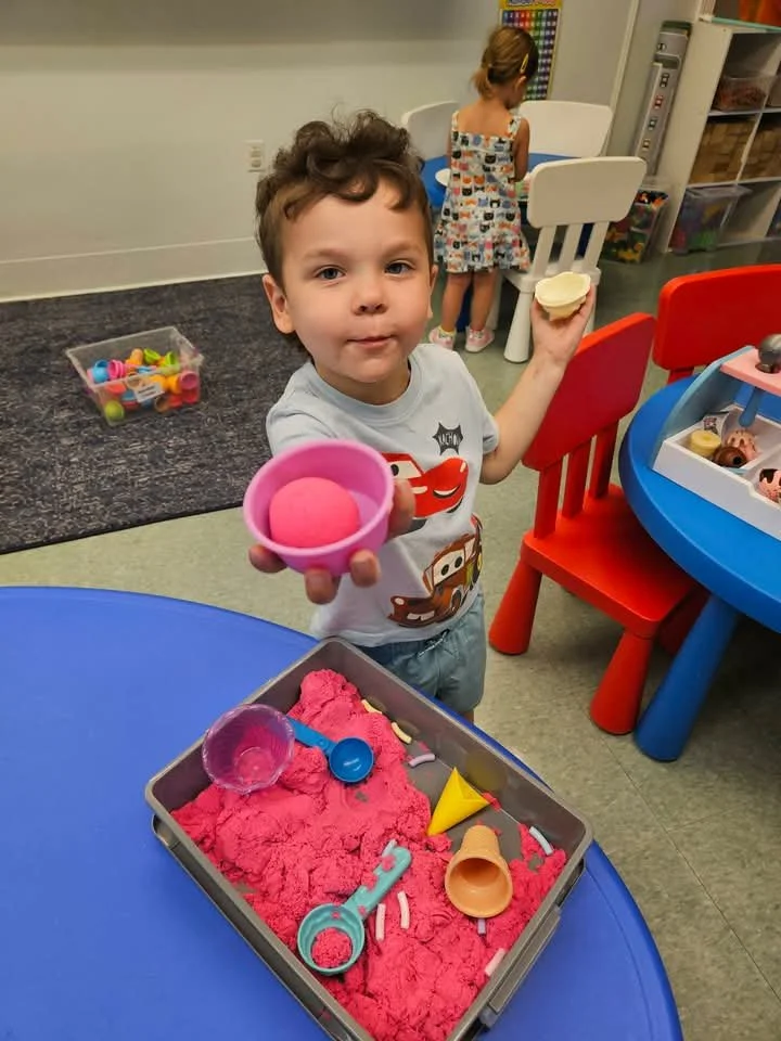 A young boy at a play area holding a pink bowl with a pink scoop and a yellow cone, standing next to a blue table with playdough and various toy utensils. In the background, a girl in a dress is facing away near a small white chair.