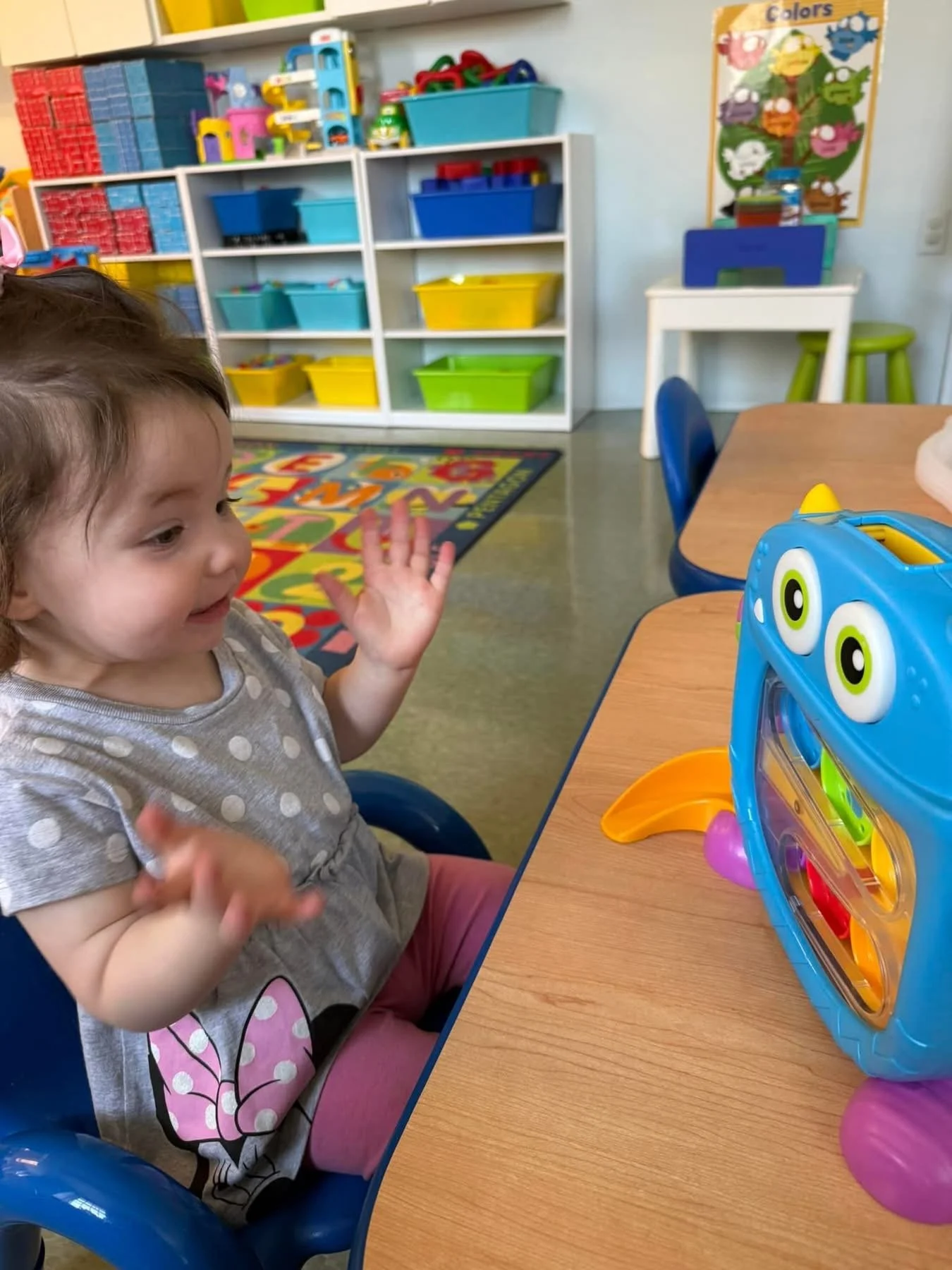 A young girl in a polka dot shirt and pink pants sitting in a blue chair at a kindergarten classroom table, smiling and waving. On the table, there is a colorful toy case shaped like a monster with large eyes and purple feet. In the background, shelv