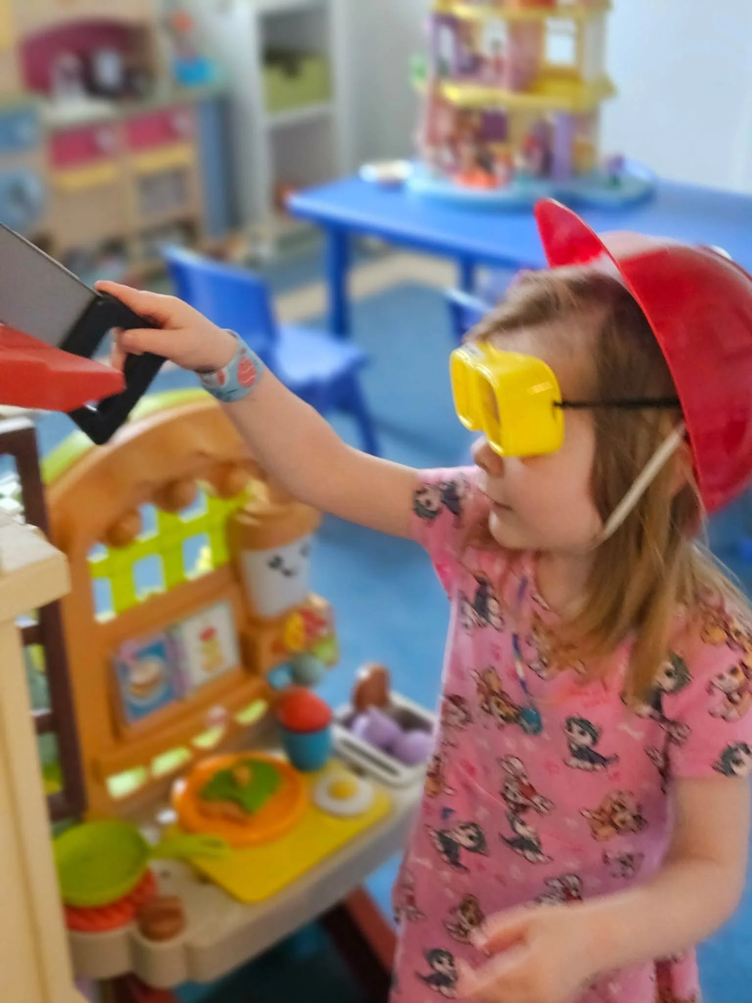 A young girl with glasses, wearing a pink shirt with cartoon characters, a red cape and yellow safety goggles, playing at a toy kitchen set in a classroom.