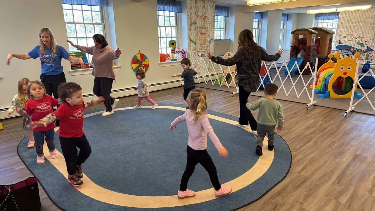 Children and adults participating in a dance or movement activity in a brightly lit indoor playroom or classroom with windows, colorful toys, and a play structure in the background.