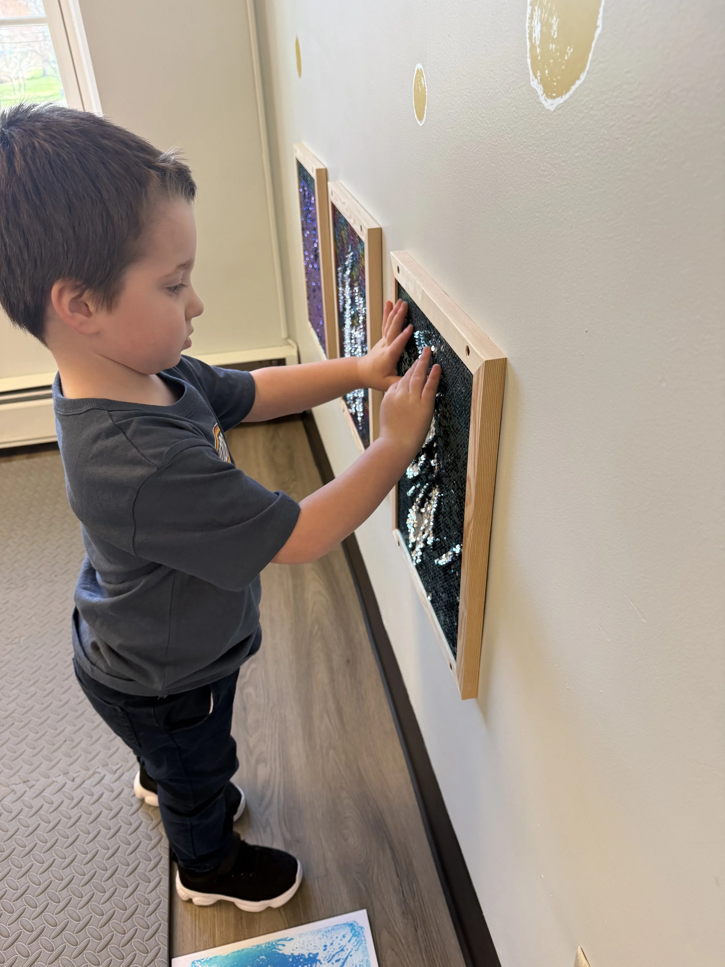 Young boy observing a black glittery art piece with sequins on a wooden frame, displayed on a beige wall.