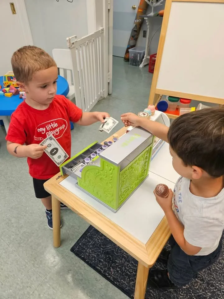 Two young boys are at a small wooden table in a classroom or daycare setting. One boy, wearing a red shirt, is handing money to the other boy, who is holding a toy cupcake. There are colorful toy items and a whiteboard in the background.