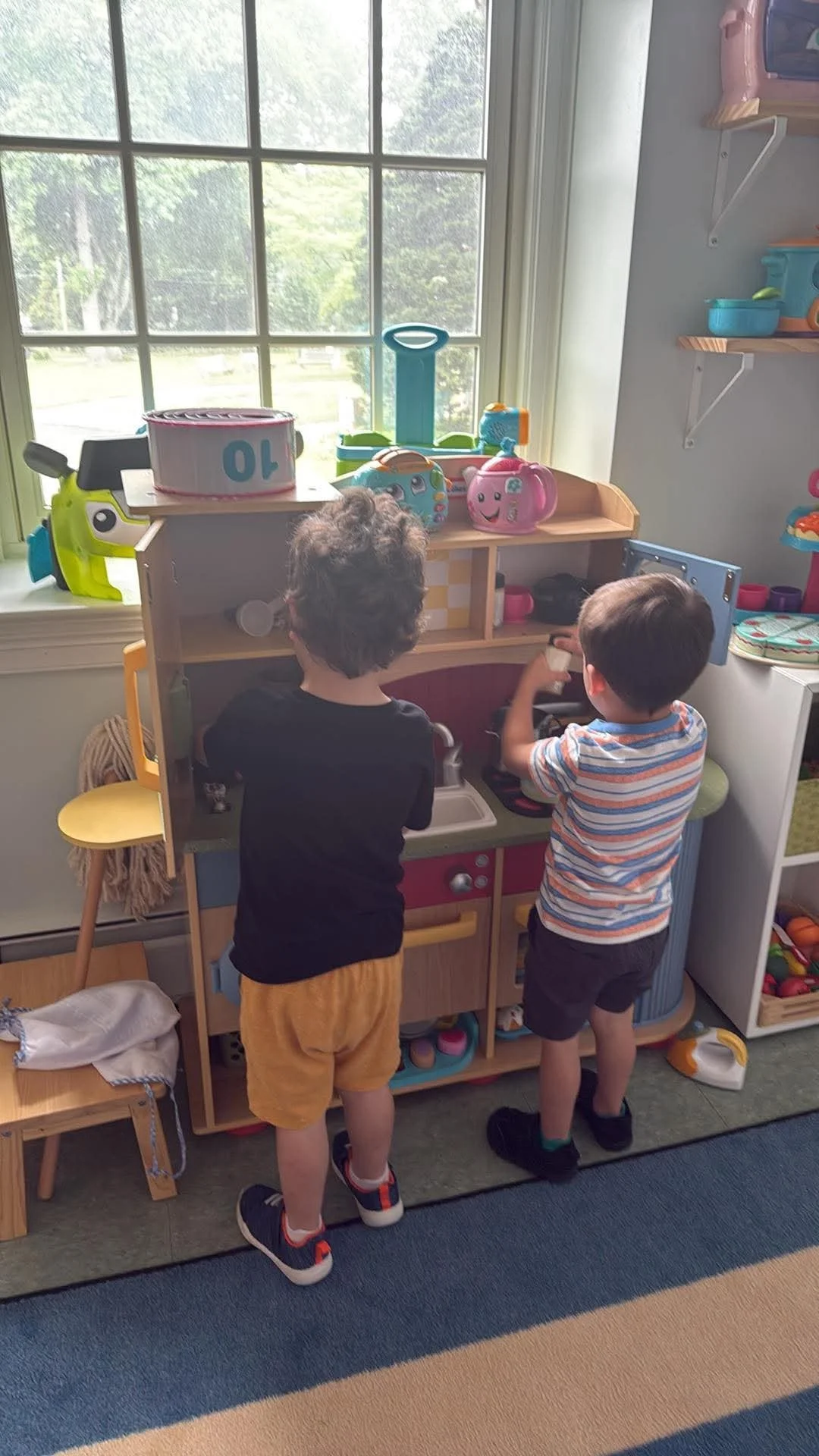 Two young boys playing with a toy kitchen set near a window, with snacks and toys on shelves around them.