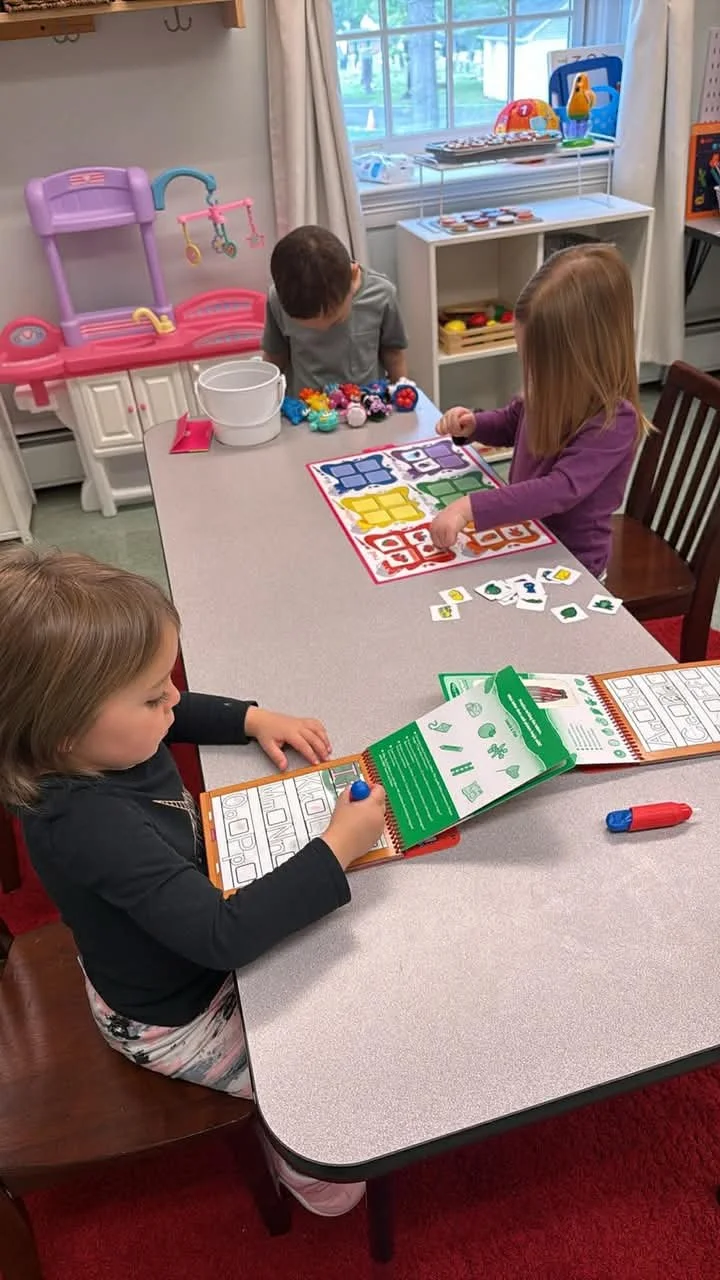 Children playing educational board games and with toys at a classroom table, with a dollhouse and window in the background.