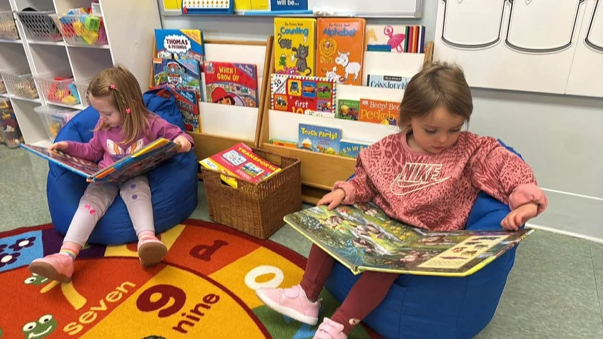 Two young girls sitting in bean bag chairs in a classroom, reading picture books. One girl is wearing a pink Nike sweatshirt and tan leggings, while the other girl is wearing a pink hoodie and patterned leggings. Behind them are shelves with children