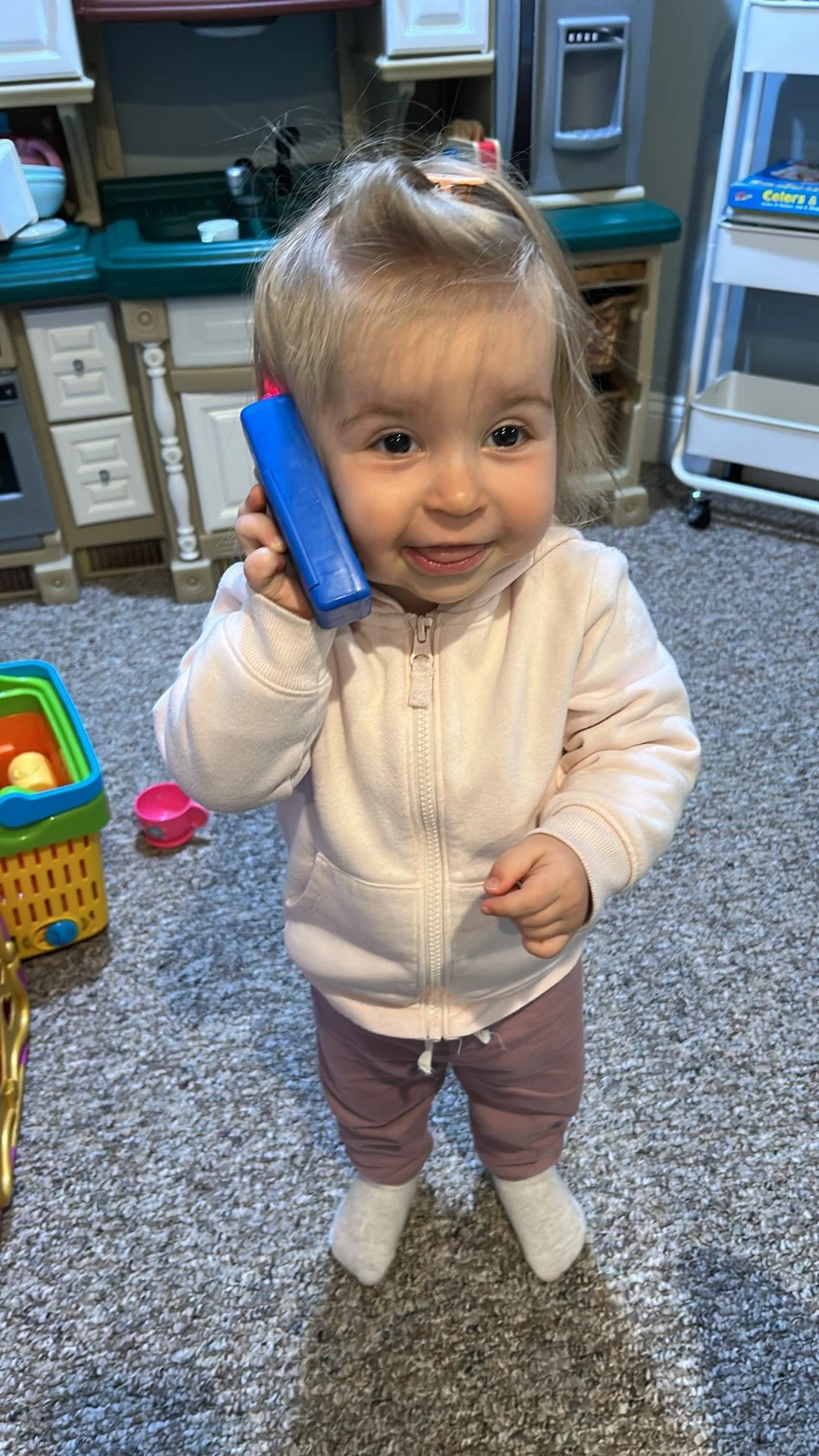 A young child with blonde hair, wearing a cream-colored zip-up hoodie, light brown pants, and socks, is standing on a carpeted floor, holding a blue toy phone to their ear, smiling.