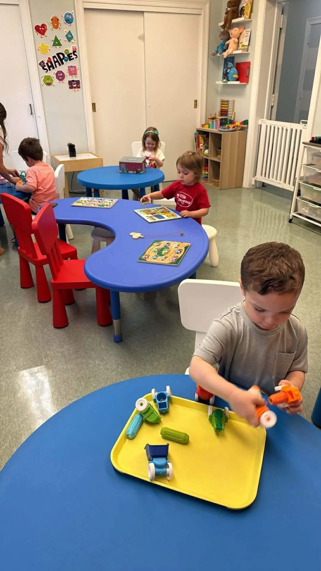 Children playing with toys in a classroom with colorful tables and chairs, shelves with books and stuffed animals, and wall decorations.