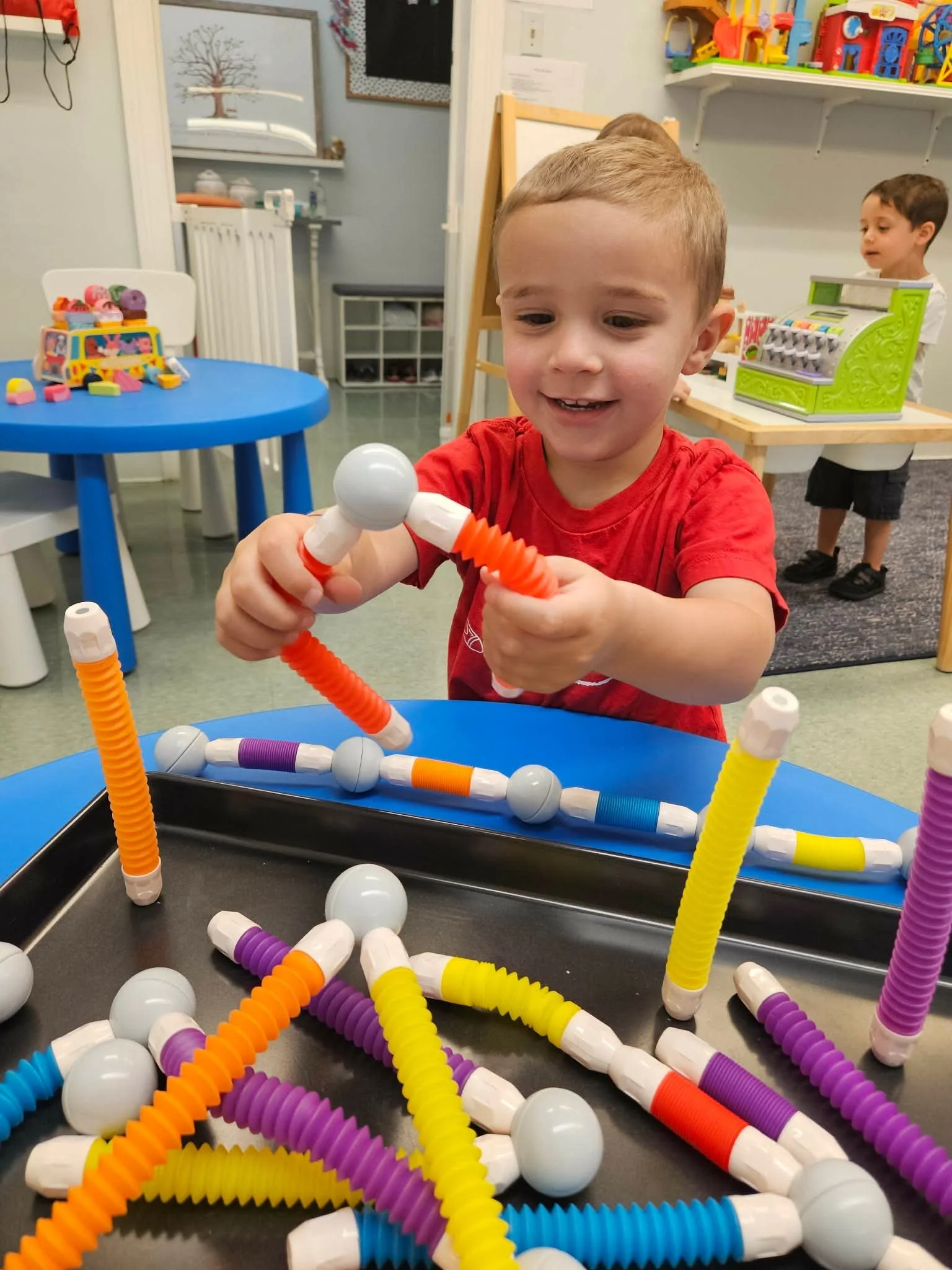 Smiling young boy in a red shirt playing with colorful building sticks and white balls at a table in a classroom or playroom.
