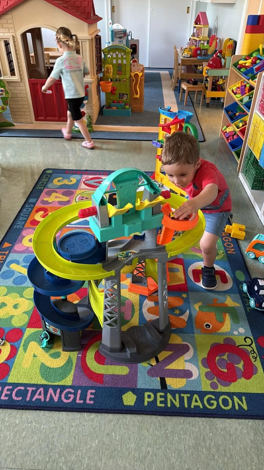 Young boy playing with a toy roller coaster in a colorful playroom, with a girl in the background near a playhouse and various toys on shelves.