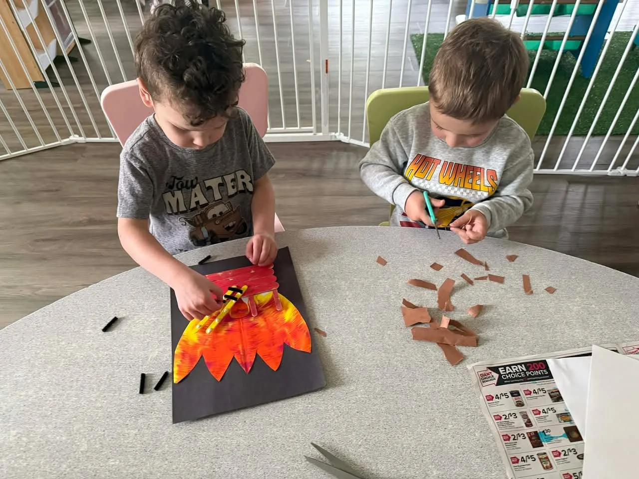 Two young boys creating artwork at a table, one working on a turkey craft and the other cutting paper, in a room with a play area in the background.
