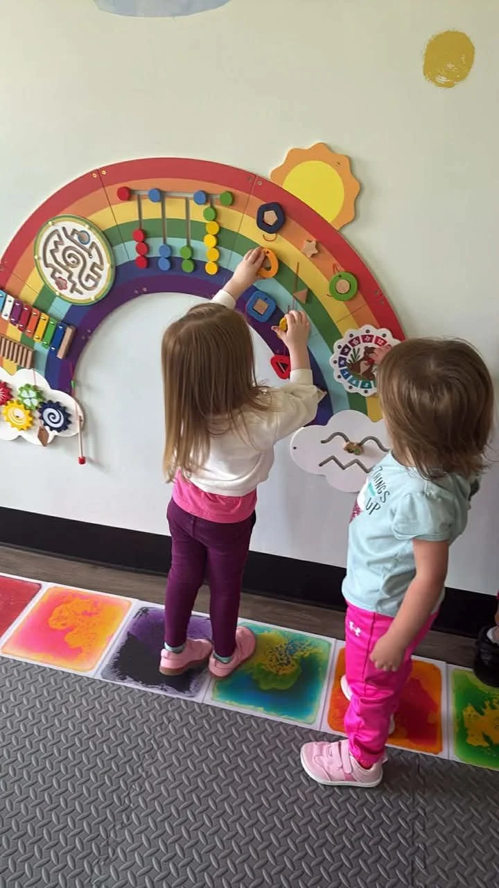 Two young children playing with a colorful educational rainbow wall hanging that has various shapes and buttons, on a carpet with bright paint splashes.