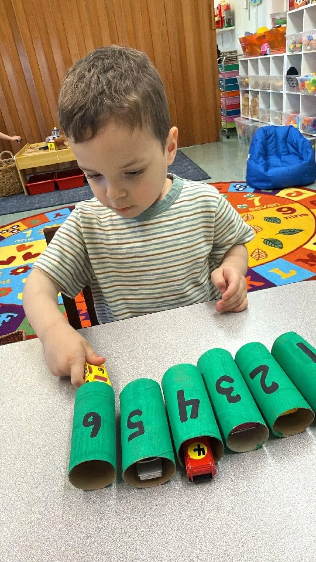 A young boy playing with green cylindrical objects labeled with numbers, in a classroom with colorful educational decor.
