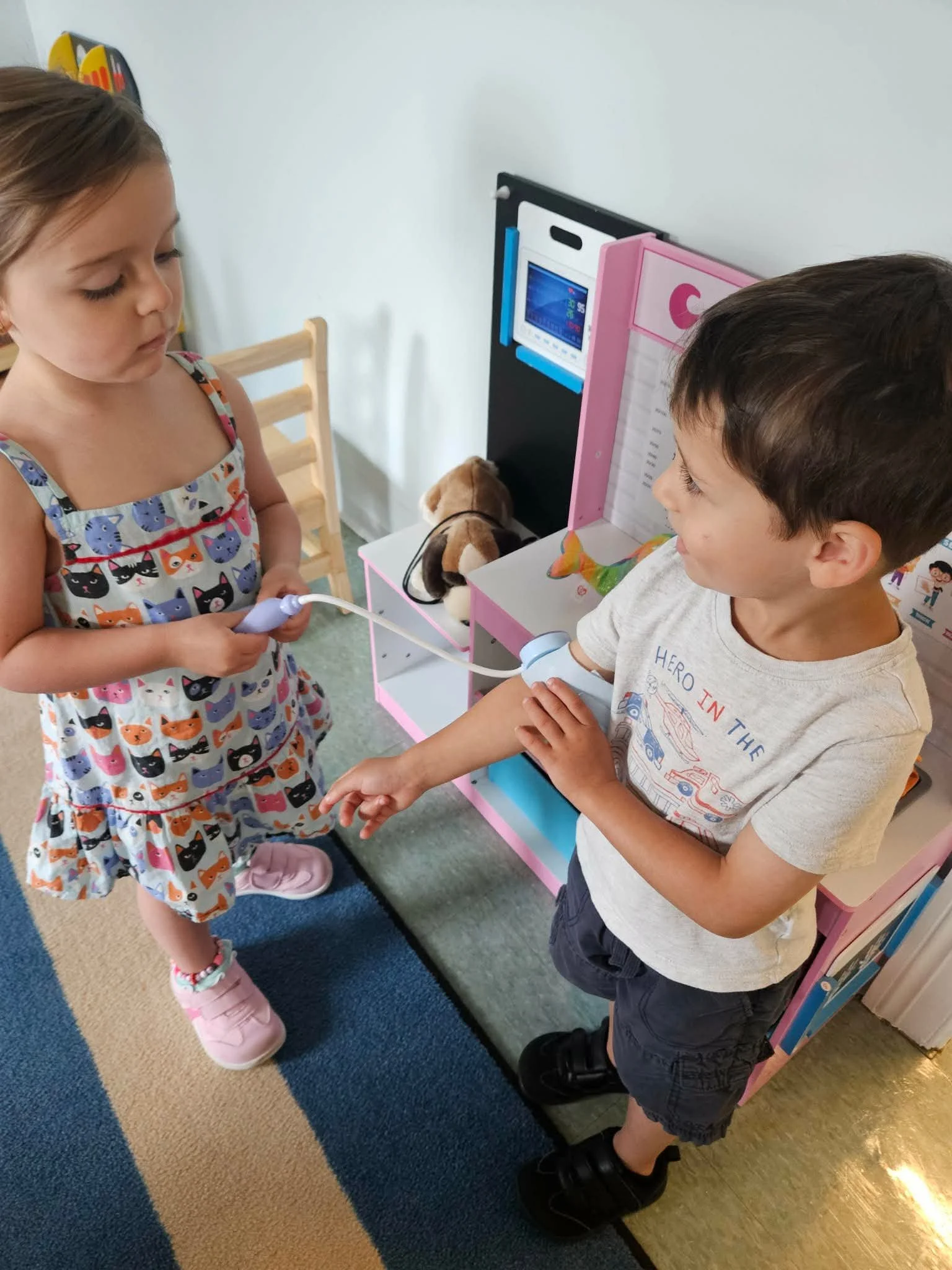 A young girl in a colorful cat-patterned dress and pink shoes transfers a liquid from a device to a boy wearing a gray t-shirt and dark shorts, in a room with a pink and black toy medical set, plush dog, and multicolored carpet.