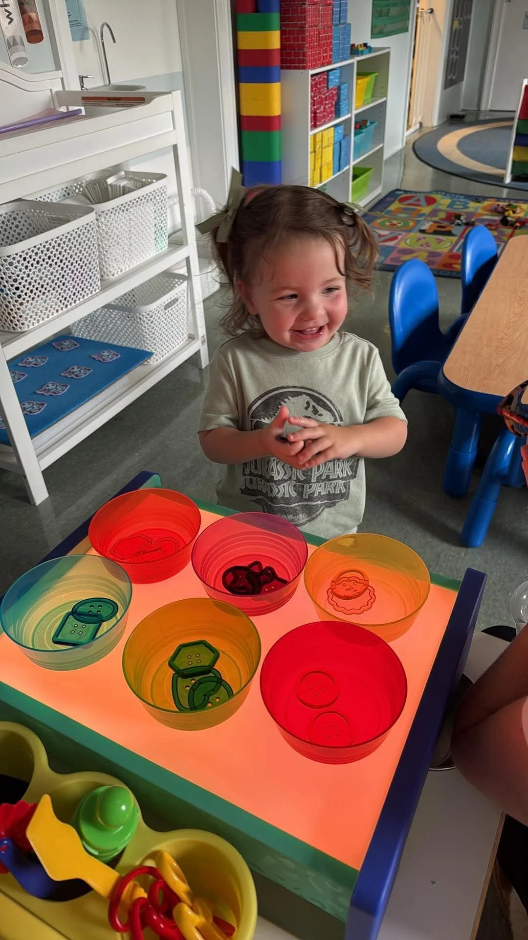 A young girl with brown hair and a bow, wearing a gray shirt, is smiling and standing at a light-up table with colorful bowls containing toy button shapes. The background shows a classroom with shelves filled with toys and educational materials, a bl
